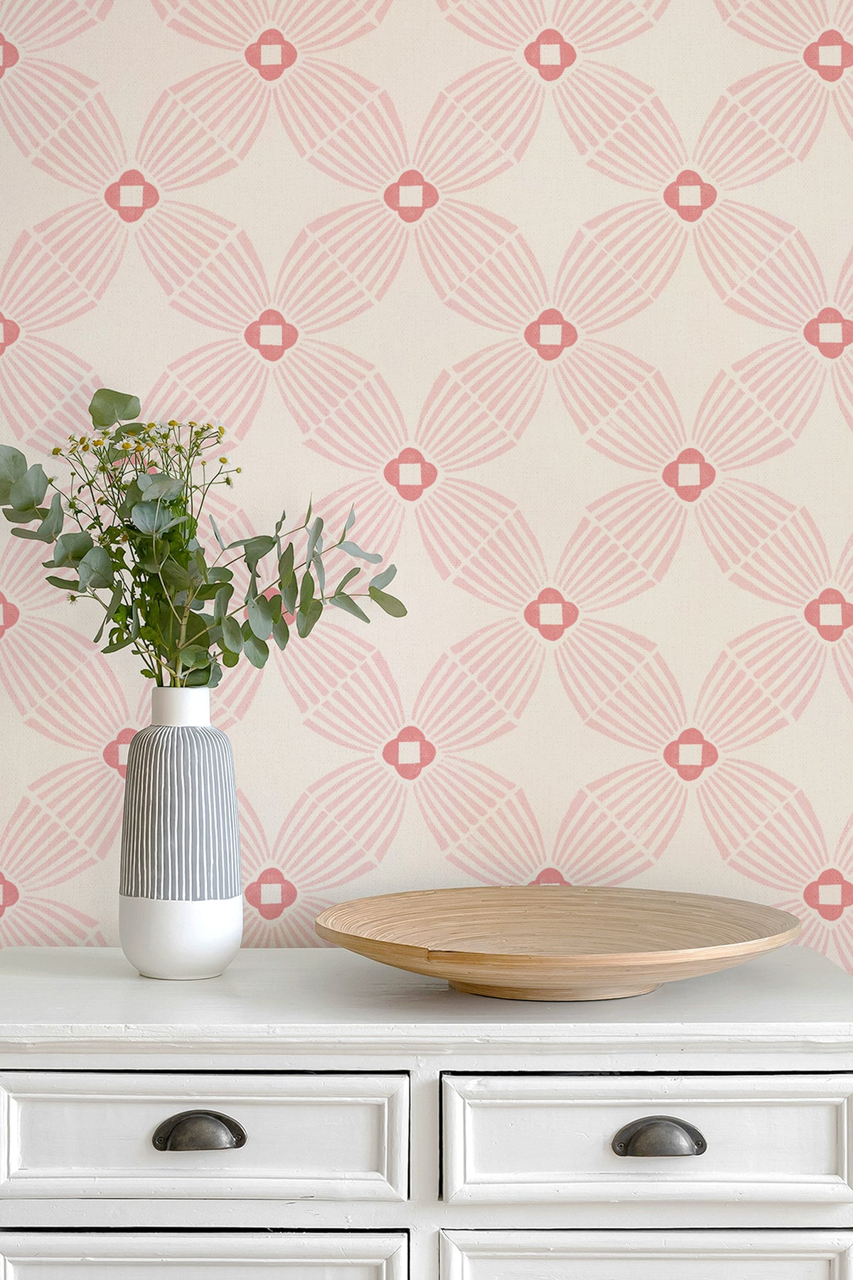 A styled interior scene showing a cream-colored wall with a repeating geometric wallpaper pattern in pink and coral, above a white dresser with a striped vase of eucalyptus and a wooden tray.