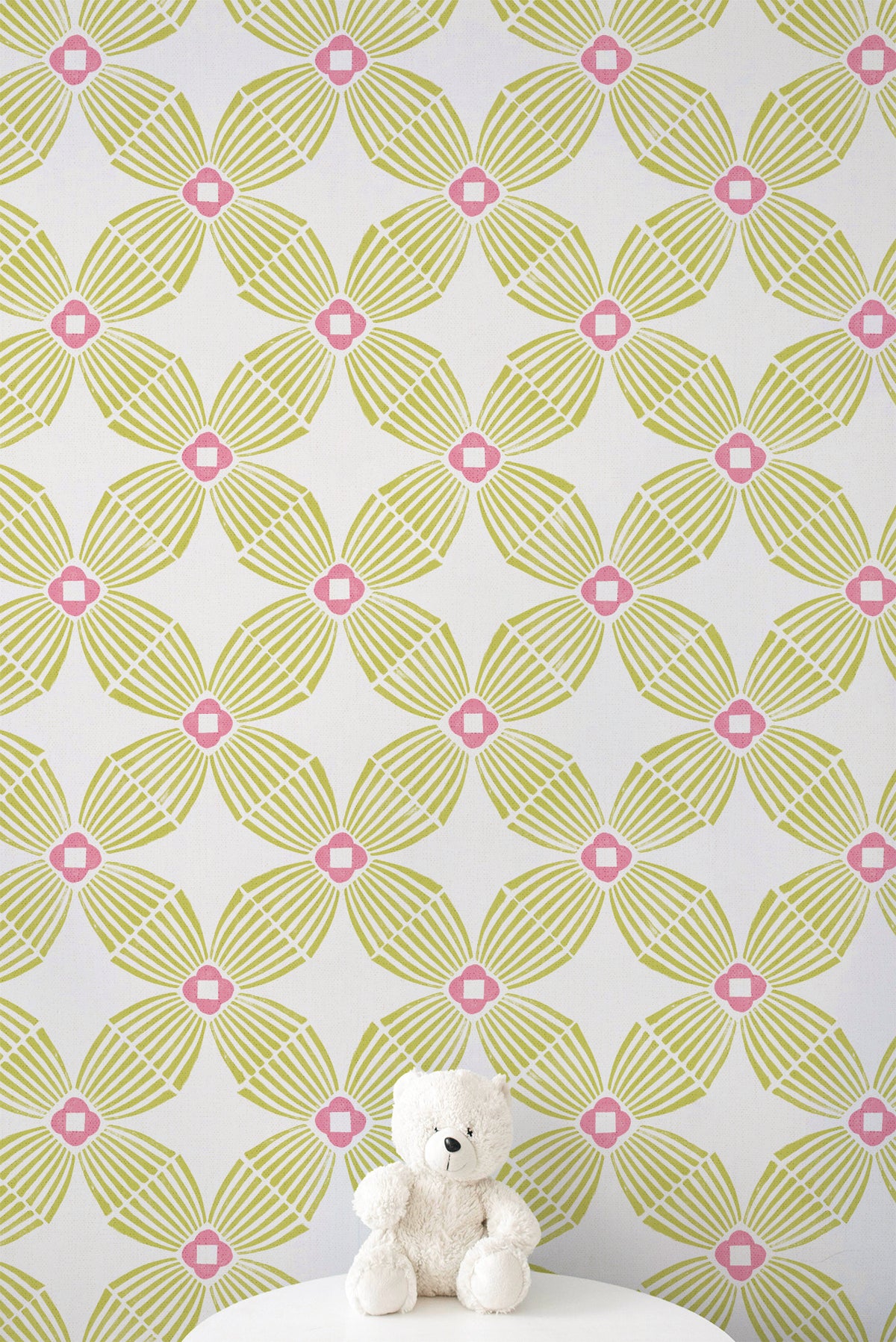 A white wall covered in a repeating geometric wallpaper pattern with lime green and pink floral motifs, with a small white teddy bear sitting on a white surface at the bottom.