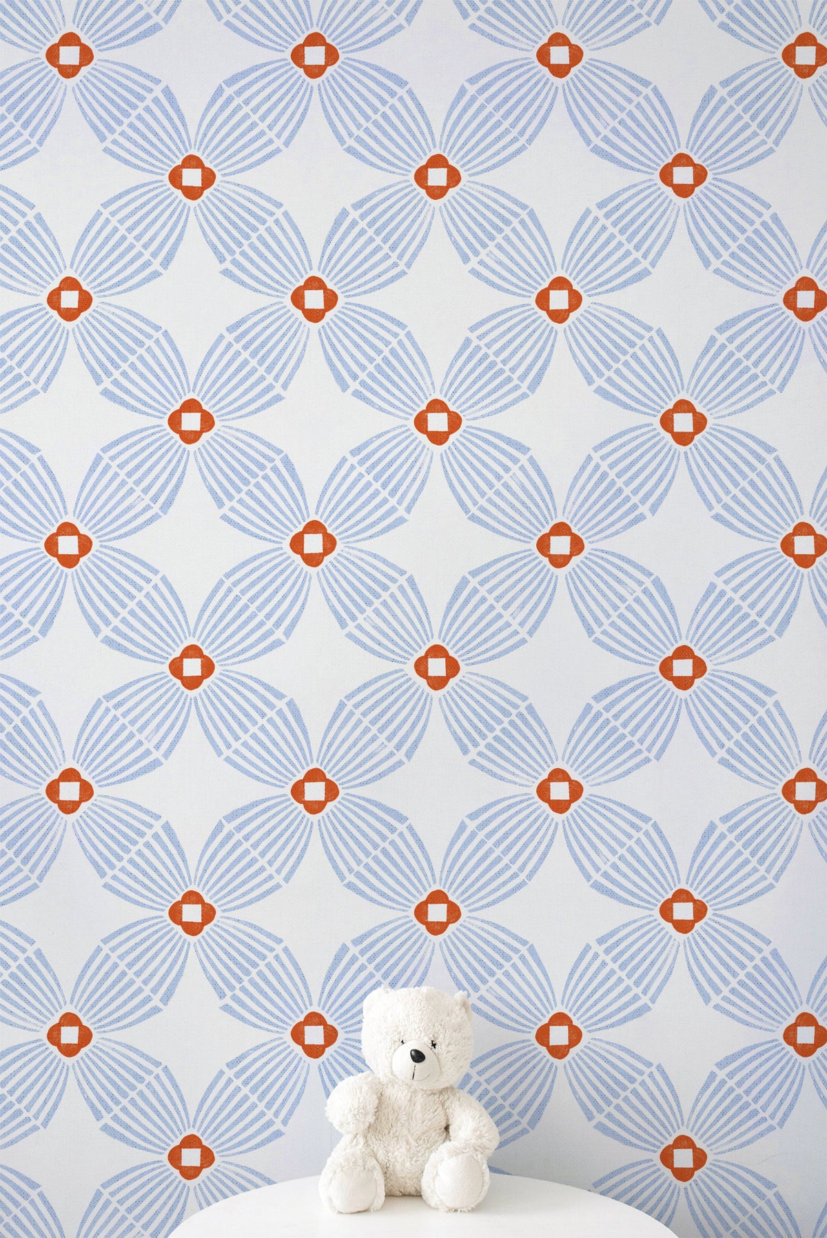 A white teddy bear sits on a white round table against a wall covered in a repeating geometric wallpaper pattern of light blue and orange.