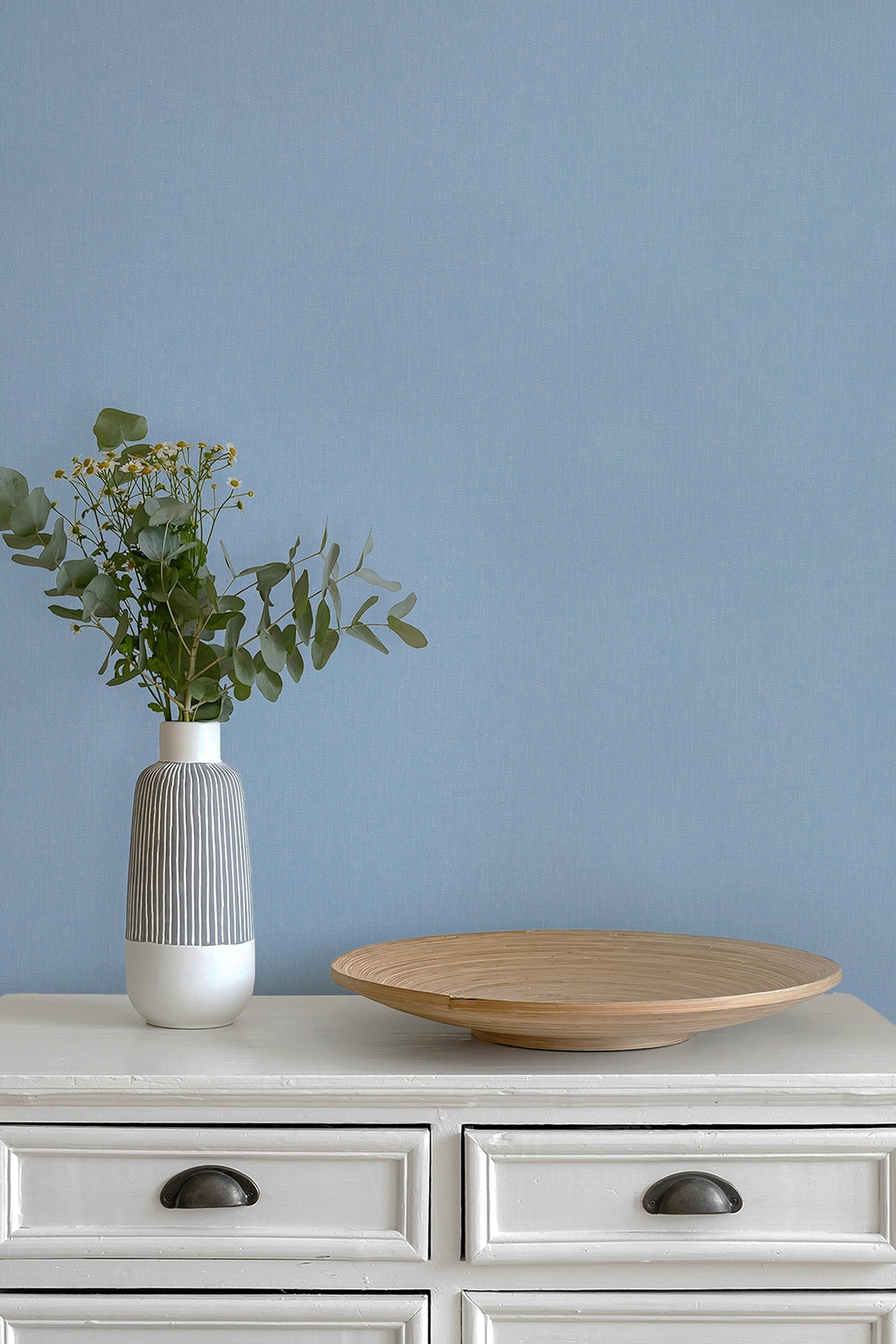 A modern gray and white ribbed ceramic vase with green eucalyptus stems, placed on a white wooden dresser next to a round light wood tray against a paperhaze wallpapered wall in periwinkle blue.