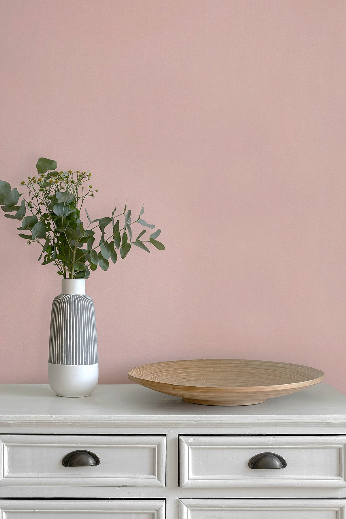 A styled interior scene with a shell pink paperhaze wallpapered wall, a white cabinet, a ribbed white and gray vase with green eucalyptus stems, and a round light wood tray.