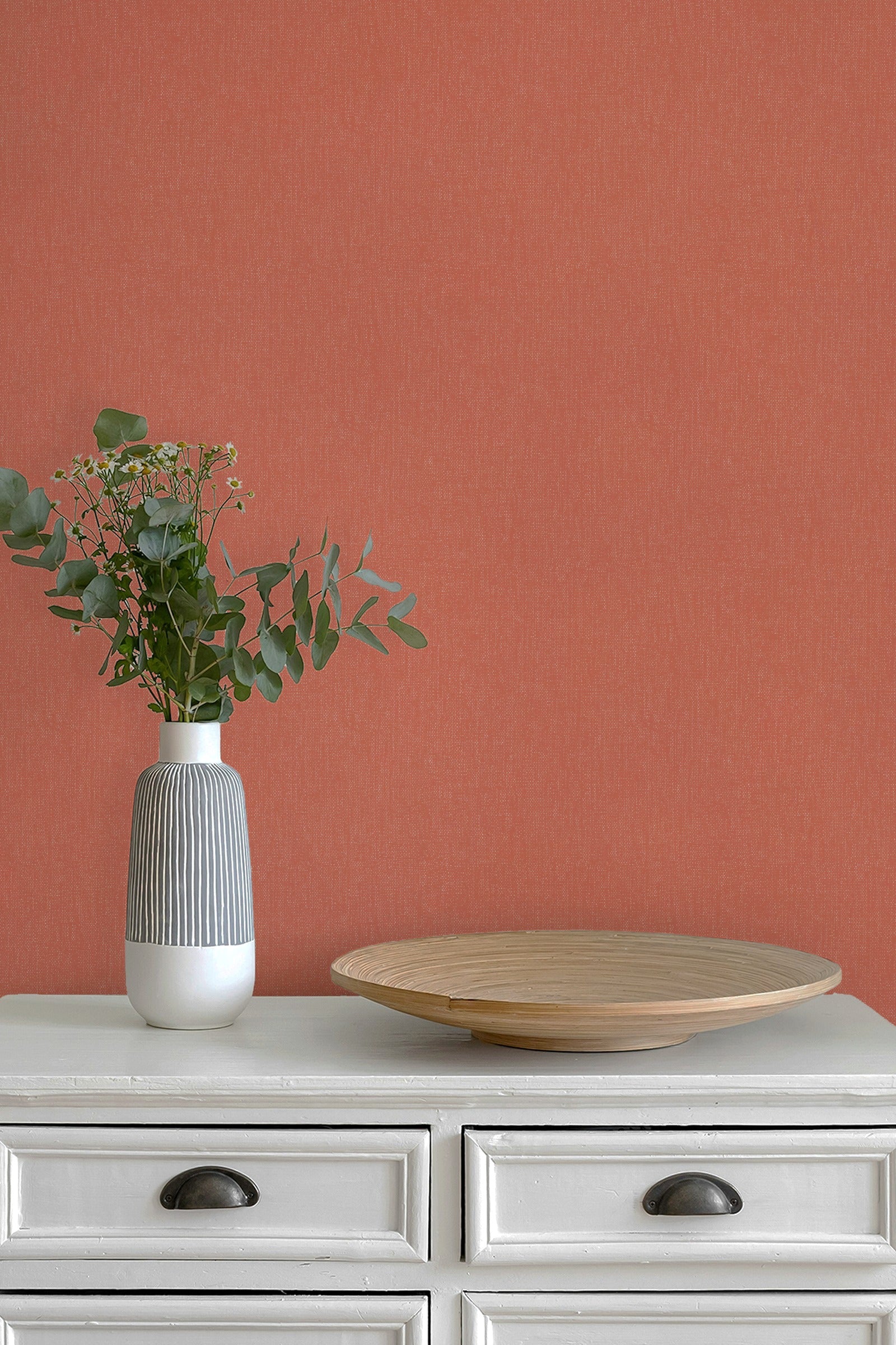 A terracotta orange textured wallpaper shown on a wall behind a white wooden dresser with a gray and white ribbed vase holding green eucalyptus stems and a round wooden tray.