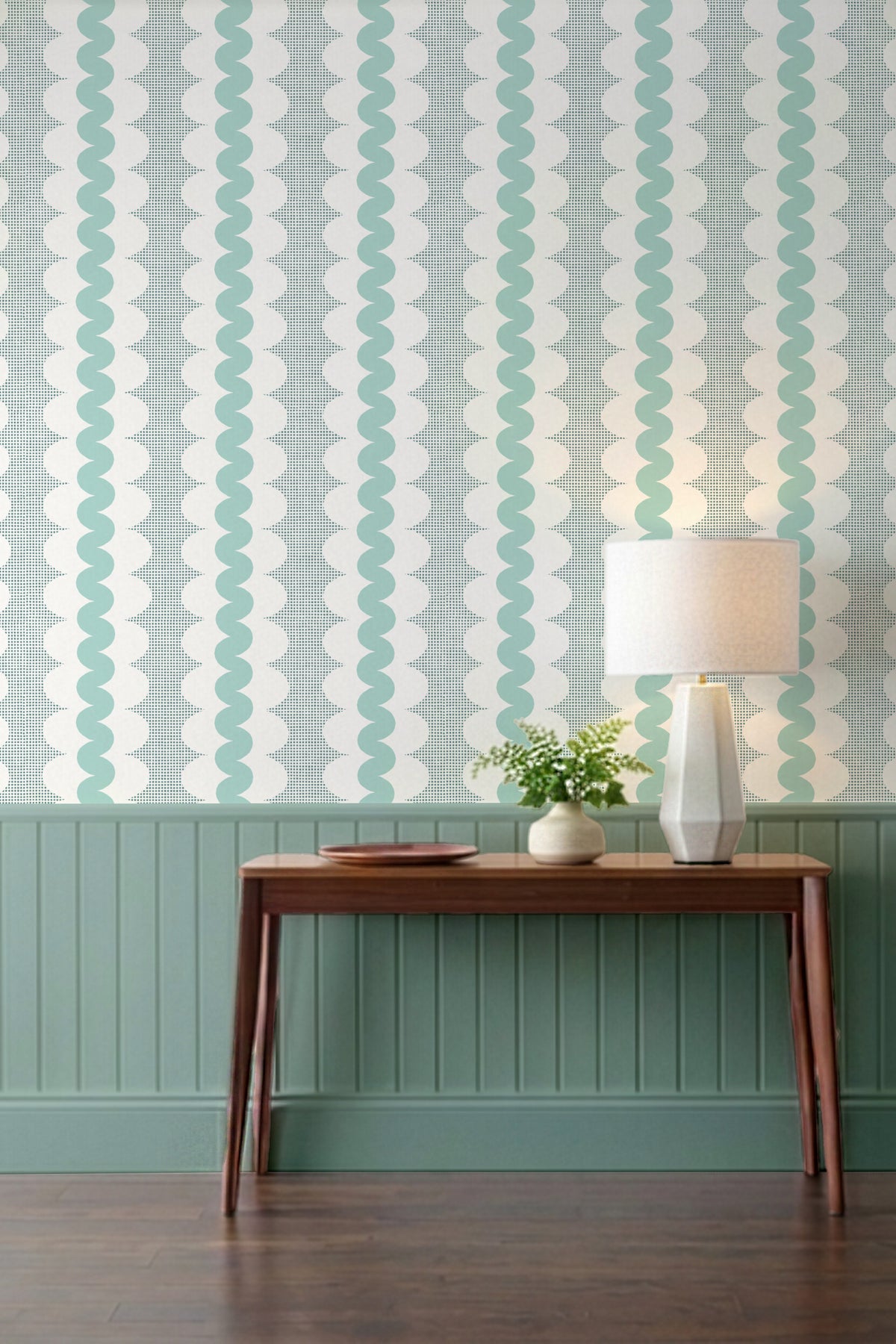 A styled interior scene showing a wall with vertical wavy stripes in mint green and white, above a teal wainscoted panel and dark wood console table. A white table lamp and a small potted plant sit on the table.
