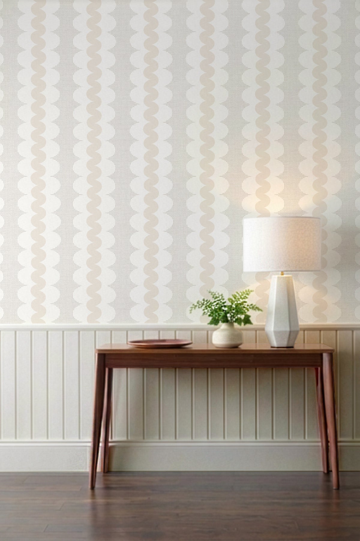 A styled interior scene showing a wall with vertical wavy patterned wallpaper, a wooden console table, a lit table lamp, and a small potted plant against a white wainscoted wall.