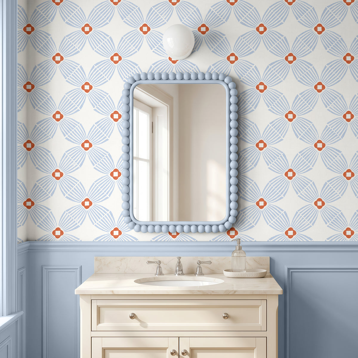 A bathroom vanity scene with a cream-colored sink cabinet, a light blue beaded mirror frame, and wallpaper featuring a repeating blue and orange floral pattern above a light blue wainscoted wall.