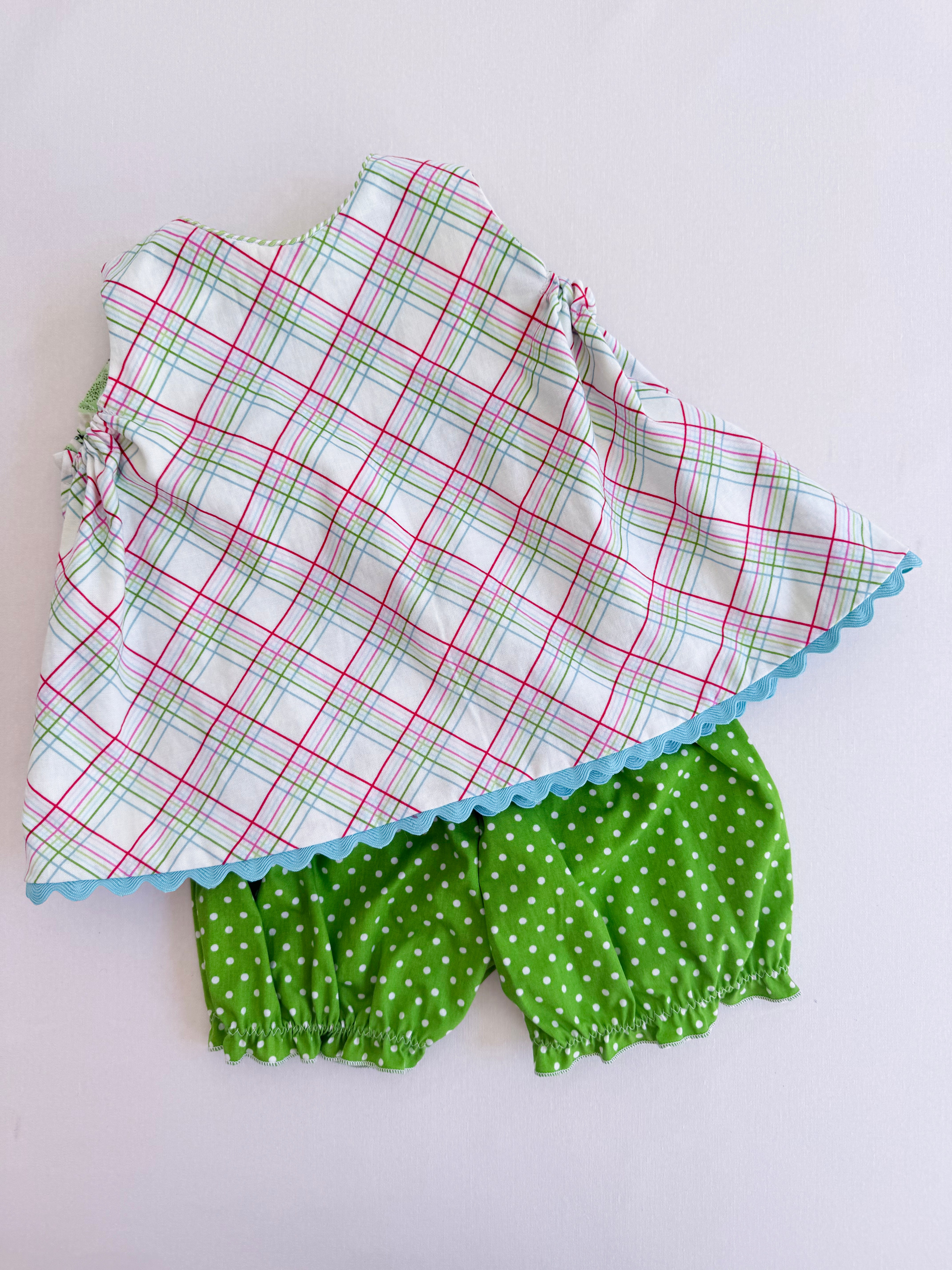A two-piece baby outfit set featuring a white sleeveless top with a red and green plaid pattern and a light blue scalloped hem, paired with green bloomers with white polka dots, laid flat on a white background.