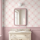 A stylized bathroom with a pink beaded mirror with a scalloped pink and white striped wallpaper background, mounted above a cream-colored vanity with a marble countertop.