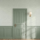 A classic six-panel interior door in a muted sage green finish, set within a matching wainscoted wall and textured wallpaper. A brass wall sconce with a white shade is mounted to the right, casting soft shadows across the lower paneling and wood floor.