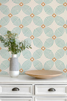 A styled interior scene showing a cream-colored wall with a repeating geometric wallpaper pattern in aqua green and warm tan, above a white dresser with a striped vase of eucalyptus and a wooden tray.