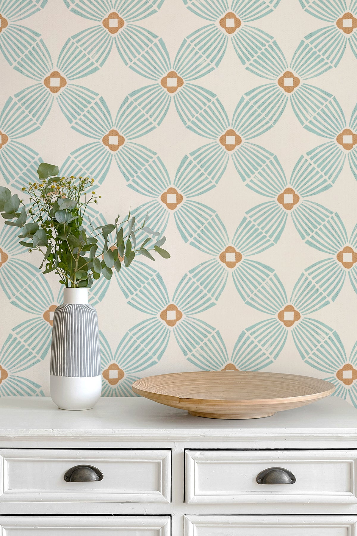 A styled interior scene showing a cream-colored wall with a repeating geometric wallpaper pattern in aqua green and warm tan, above a white dresser with a striped vase of eucalyptus and a wooden tray.
