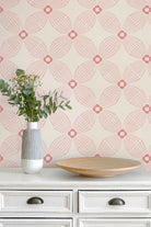 A styled interior scene showing a cream-colored wall with a repeating geometric wallpaper pattern in pink and coral, above a white dresser with a striped vase of eucalyptus and a wooden tray.