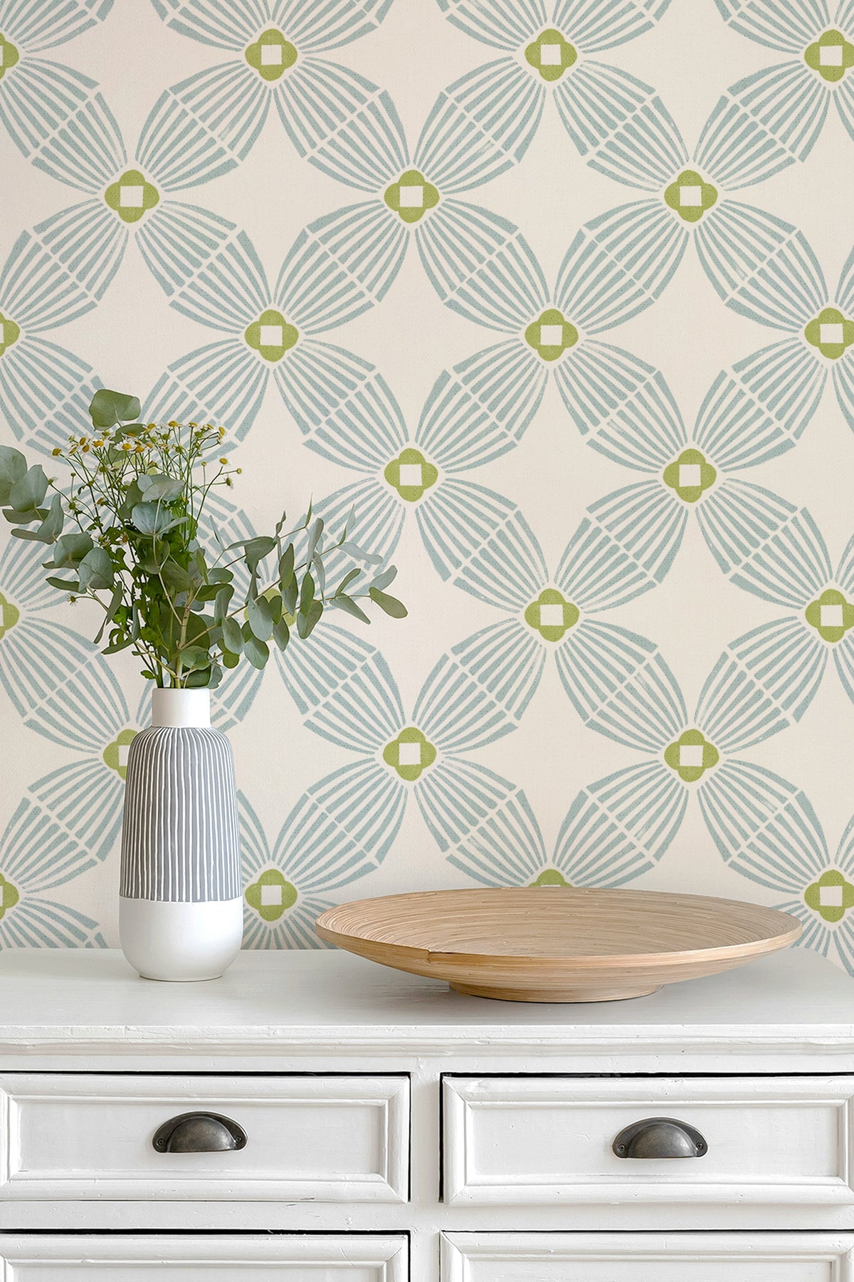 A styled interior scene showing a cream-colored wall with a repeating geometric wallpaper pattern in aqua green and lime, above a white dresser with a striped vase of eucalyptus and a wooden tray.