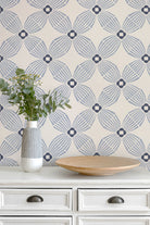 A styled interior scene showing a cream wallpaper with a repeating blue-gray geometric fan pattern on a white dresser. A white vase with green stems and a shallow round wooden tray sit on the dresser.