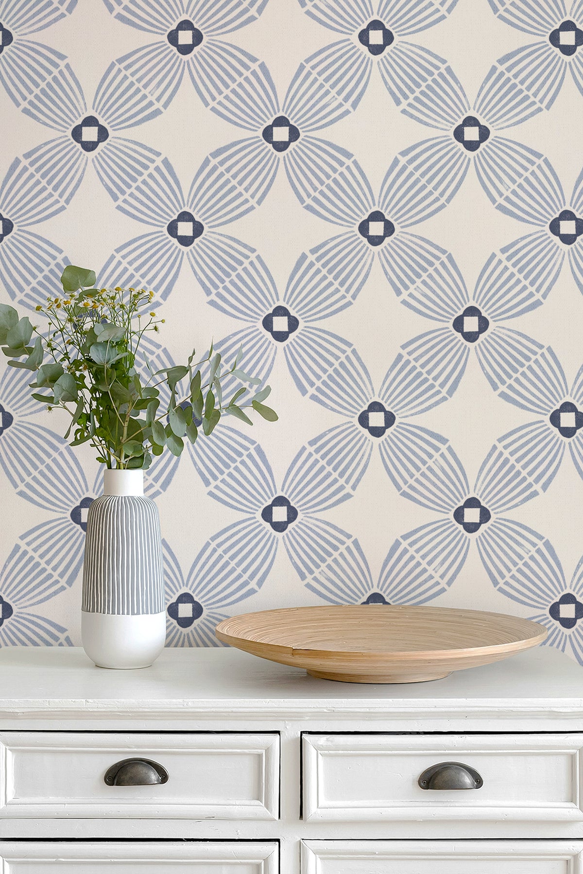 A styled interior scene showing a cream wallpaper with a repeating blue-gray geometric fan pattern on a white dresser. A white vase with green stems and a shallow round wooden tray sit on the dresser.