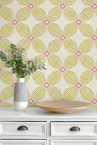 A styled interior scene showing a cream-colored wall with a repeating geometric wallpaper pattern in lime green and hot pink, above a white dresser with a striped vase of eucalyptus and a wooden tray.
