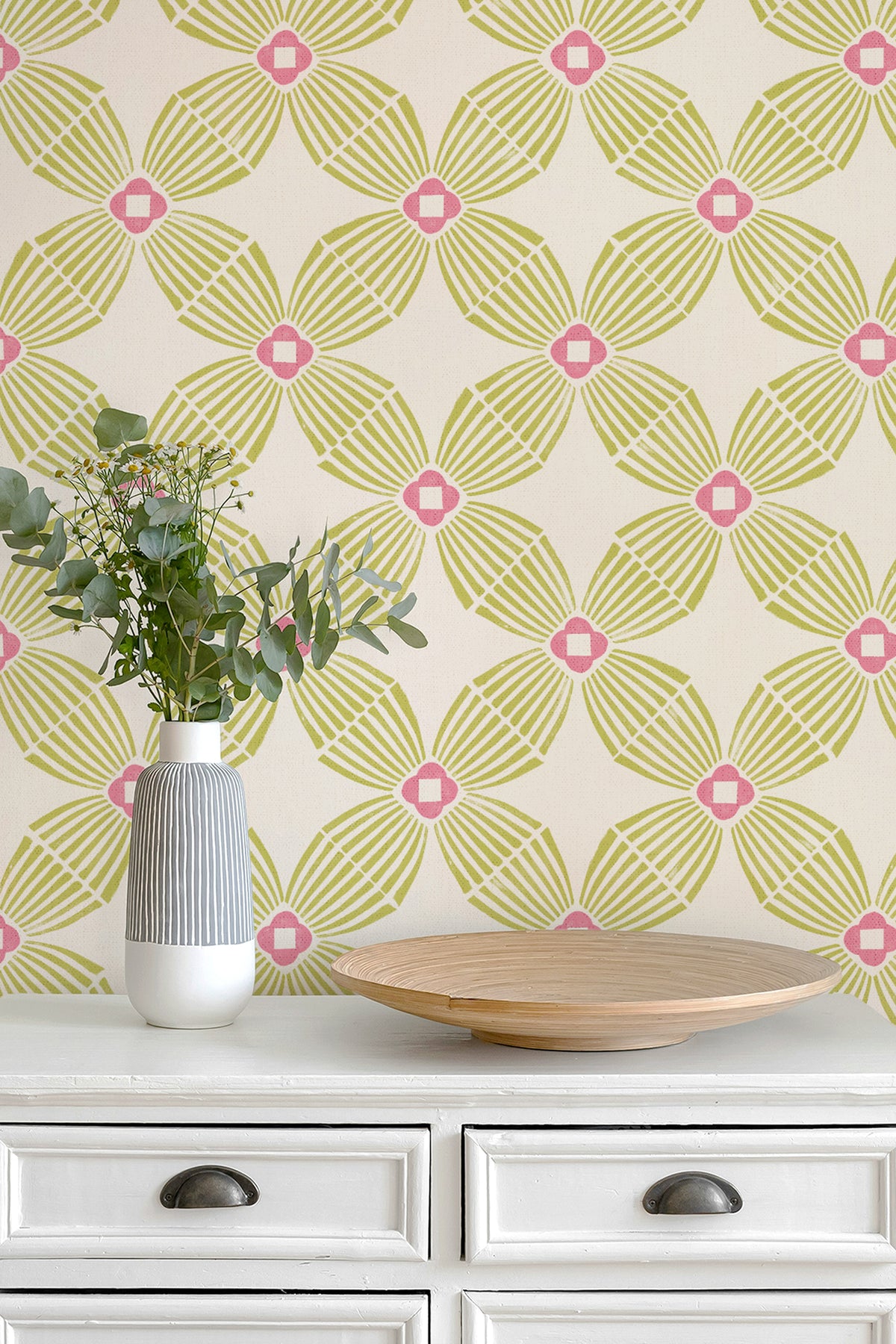 A styled interior scene showing a cream-colored wall with a repeating geometric wallpaper pattern in lime green and hot pink, above a white dresser with a striped vase of eucalyptus and a wooden tray.