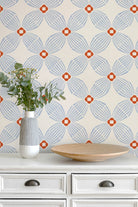 A styled interior scene showing a cream wallpaper with a repeating geometric medallion pattern in light blue and rust orange, mounted above a white dresser with a striped vase and a wooden bowl.