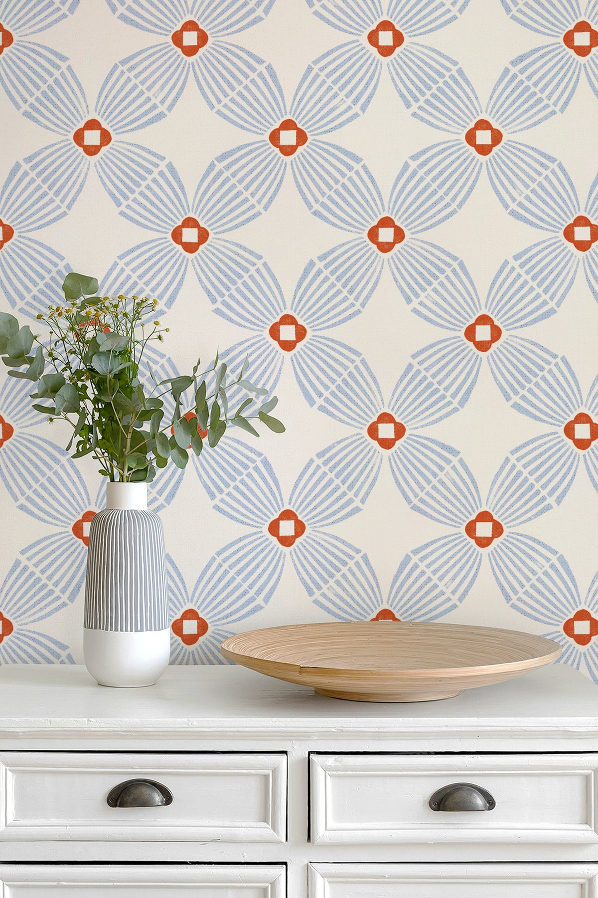 A styled interior scene showing a cream wallpaper with a repeating geometric medallion pattern in light blue and rust orange, mounted above a white dresser with a striped vase and a wooden bowl.