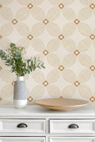 A styled interior scene showing a cream wallpaper with a repeating geometric fan pattern in warm tan, mounted above a white dresser. A white and gray ribbed vase with greenery and a round wooden tray sit on the dresser.