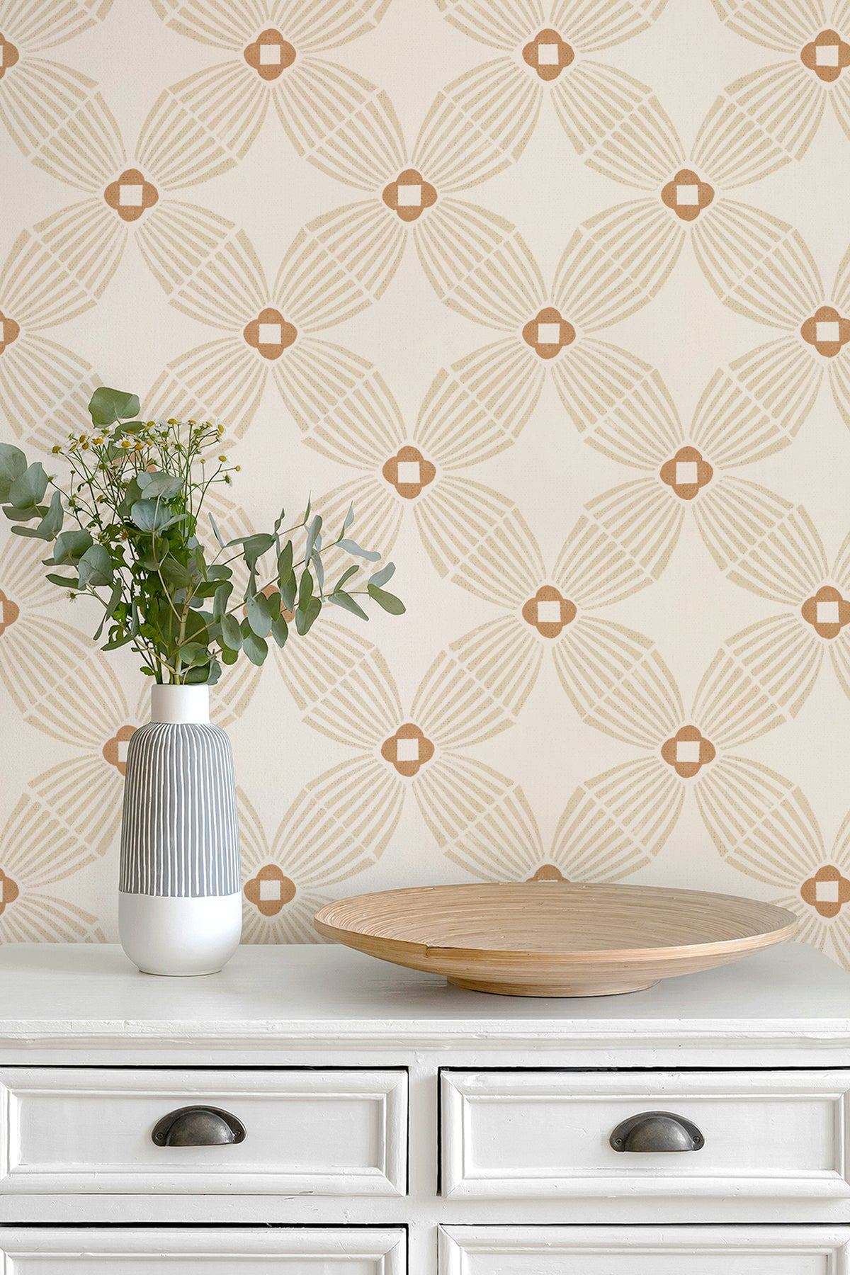 A styled interior scene showing a cream wallpaper with a repeating geometric fan pattern in warm tan, mounted above a white dresser. A white and gray ribbed vase with greenery and a round wooden tray sit on the dresser.