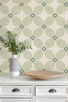 A styled interior scene showing a cream-colored wall with sage green geometric wallpaper, a white dresser, a striped ceramic vase with greenery, and a round wooden tray.