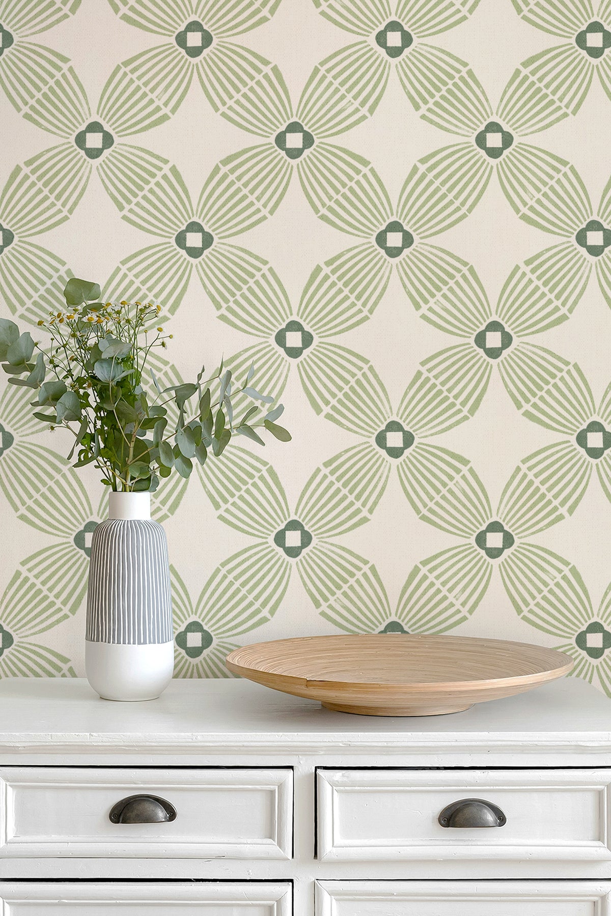 A styled interior scene showing a cream-colored wall with sage green geometric wallpaper, a white dresser, a striped ceramic vase with greenery, and a round wooden tray.