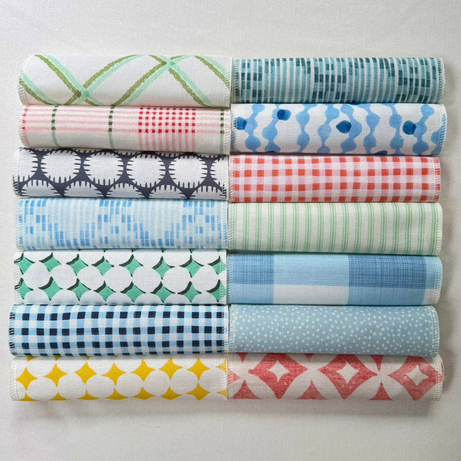 Twelve folded fabric rolls arranged in two columns of six on a light off-white background. The fabrics feature various patterns including stripes, checks, dots, and geometric shapes in shades of blue, green, pink, red, yellow, and navy.