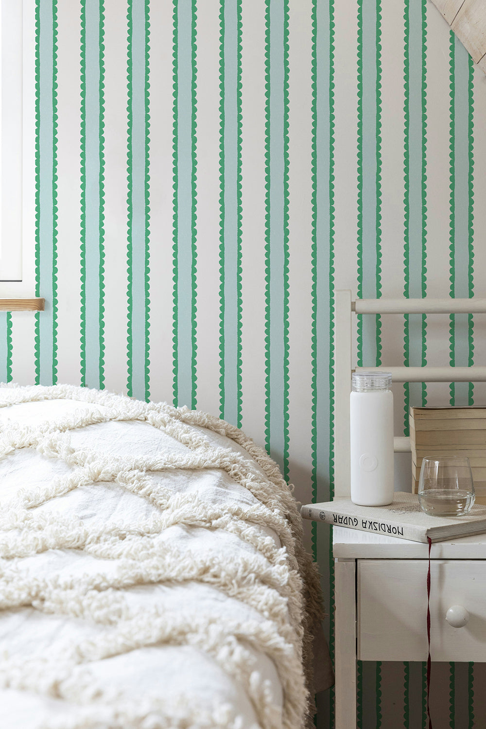 Crayon scallops in green, aqua and white vertically striped wallpaper in a bedroom setting, styled with a textured cream quilt and a white bedside table.