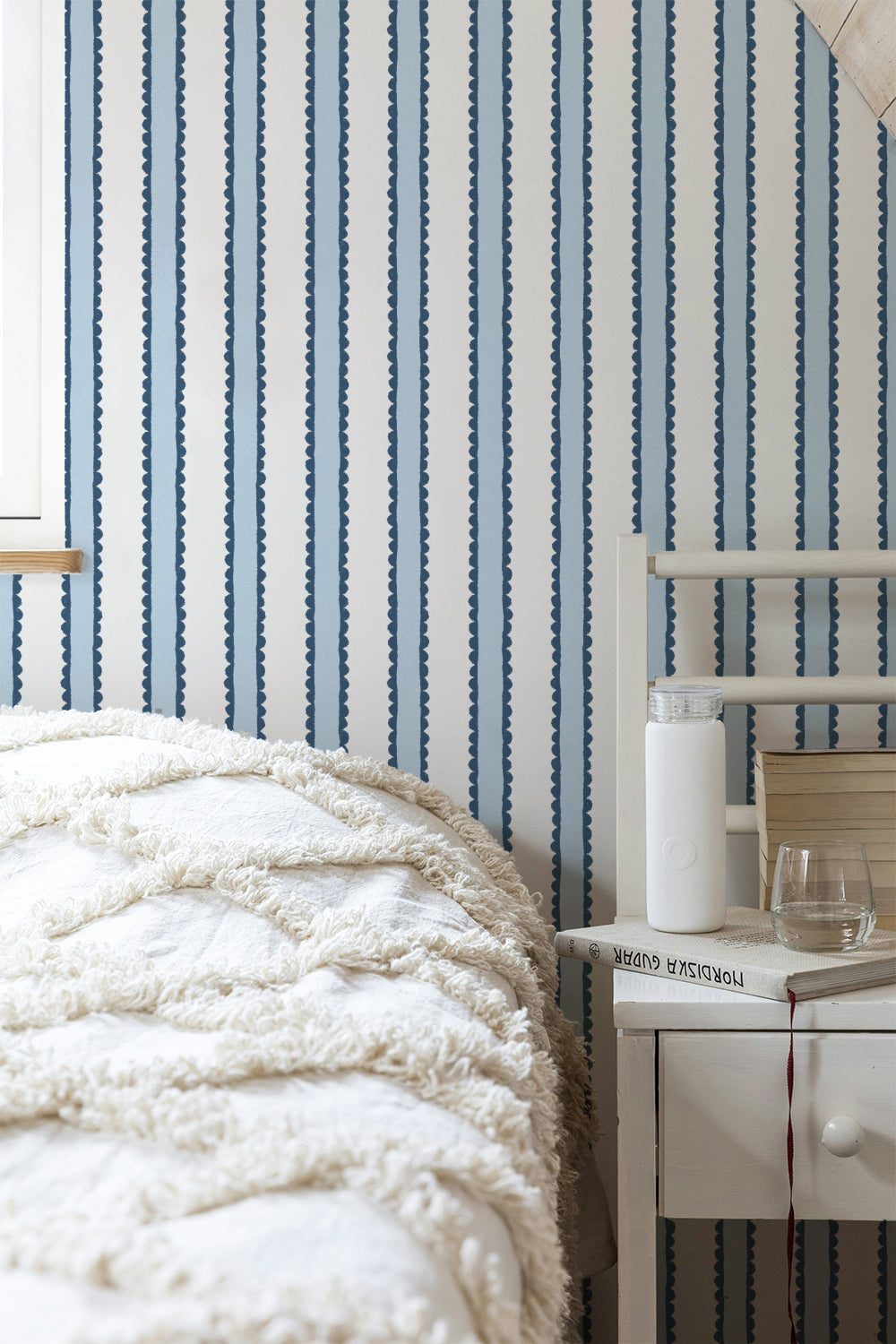Blue and white vertically striped wallpaper in a bedroom setting, shown behind a bed with a textured cream blanket and a white bedside table.