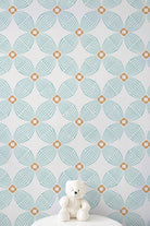 A white teddy bear sits on a white pedestal against a wall with a repeating geometric pattern in light aqua and tan.