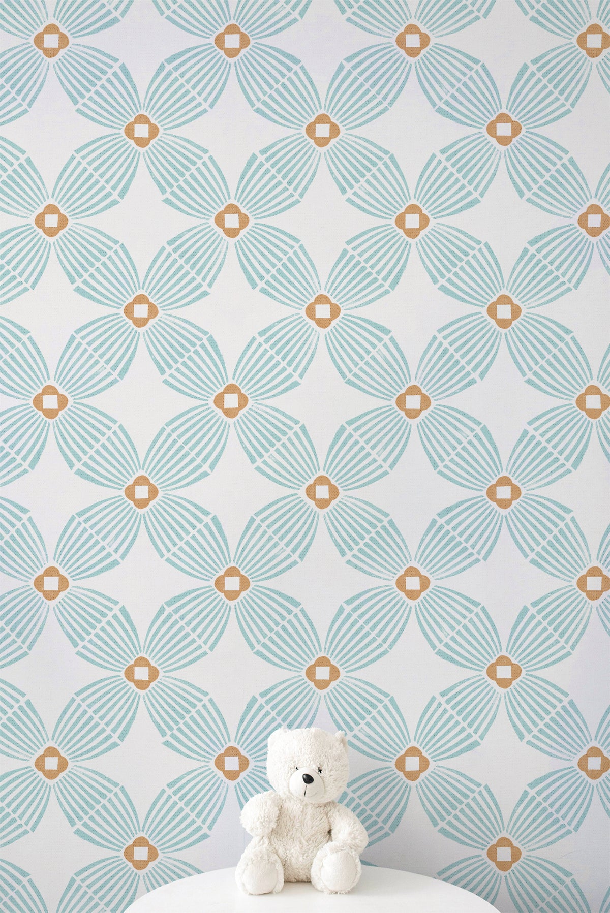 A white teddy bear sits on a white pedestal against a wall with a repeating geometric pattern in light aqua and tan.
