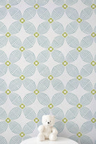 A white teddy bear sits on a white pedestal against a wall with a repeating geometric pattern in light aqua gray and lime green
