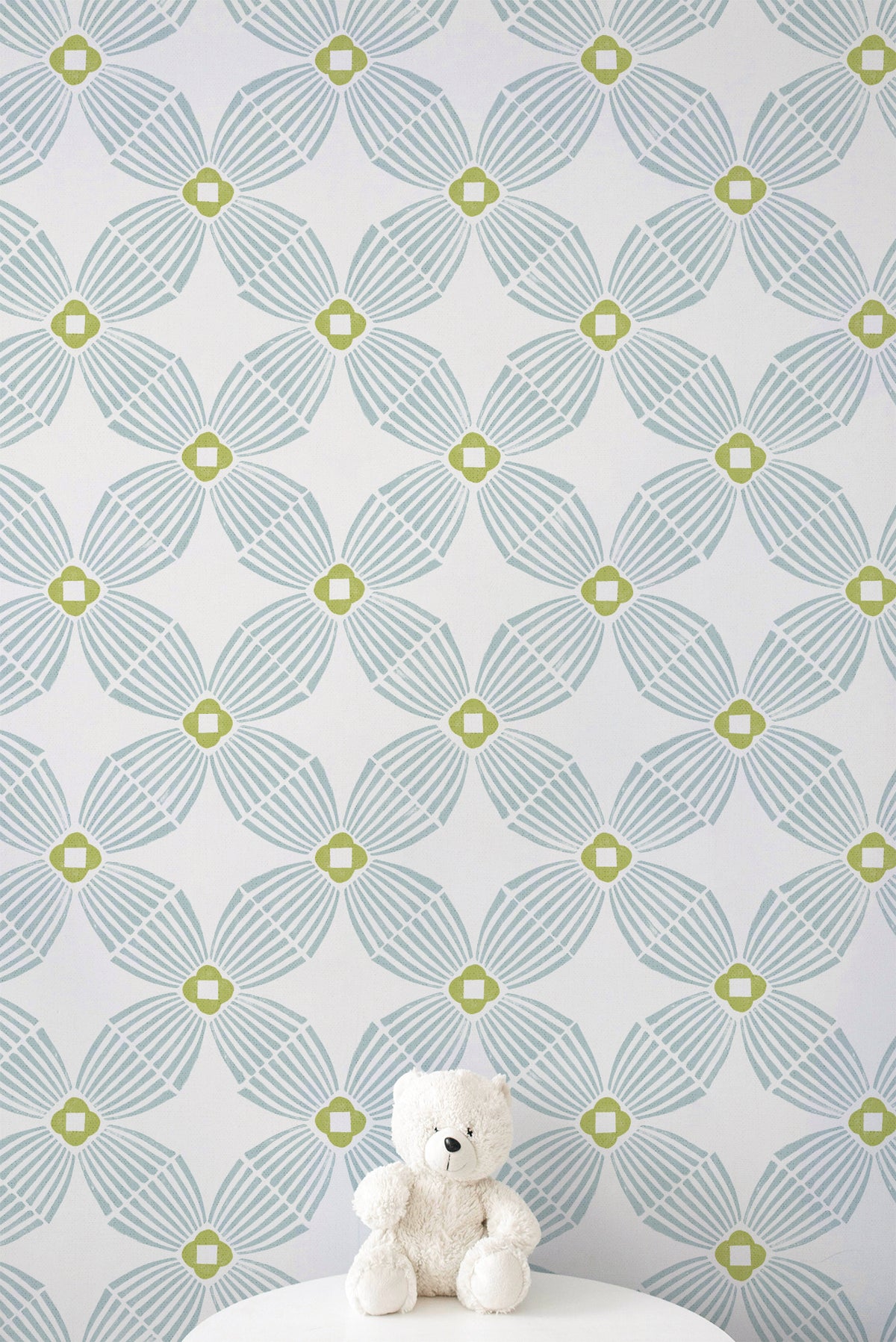 A white teddy bear sits on a white pedestal against a wall with a repeating geometric pattern in light aqua gray and lime green