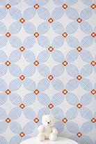 A white teddy bear sits on a white round table against a wall covered in a repeating geometric wallpaper pattern of light blue and orange.