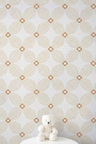 A white teddy bear sits on a white pedestal against a wall covered in repeating geometric wallpaper with a warm beige and white pattern.