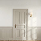 A classic six-panel interior door in a warm beige/cream finish, set within a matching door frame on a wall with paperhaze wallpaper in warm beige with white wainscoting and a gold wall sconce.