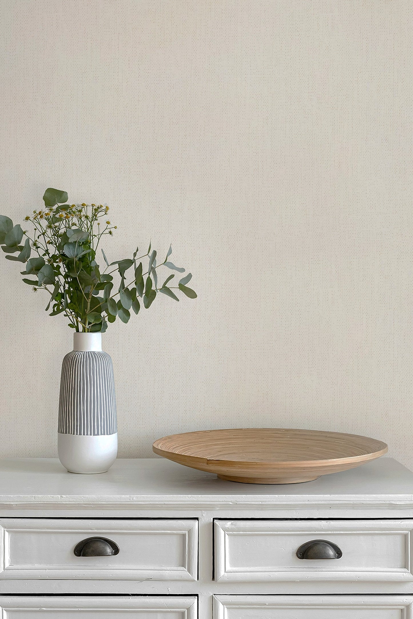A modern ceramic vase with a gray ribbed body and white base holding green eucalyptus stems, placed on a white cabinet next to a round wooden tray against a wall in paperhaze wallpaper warm beige.
