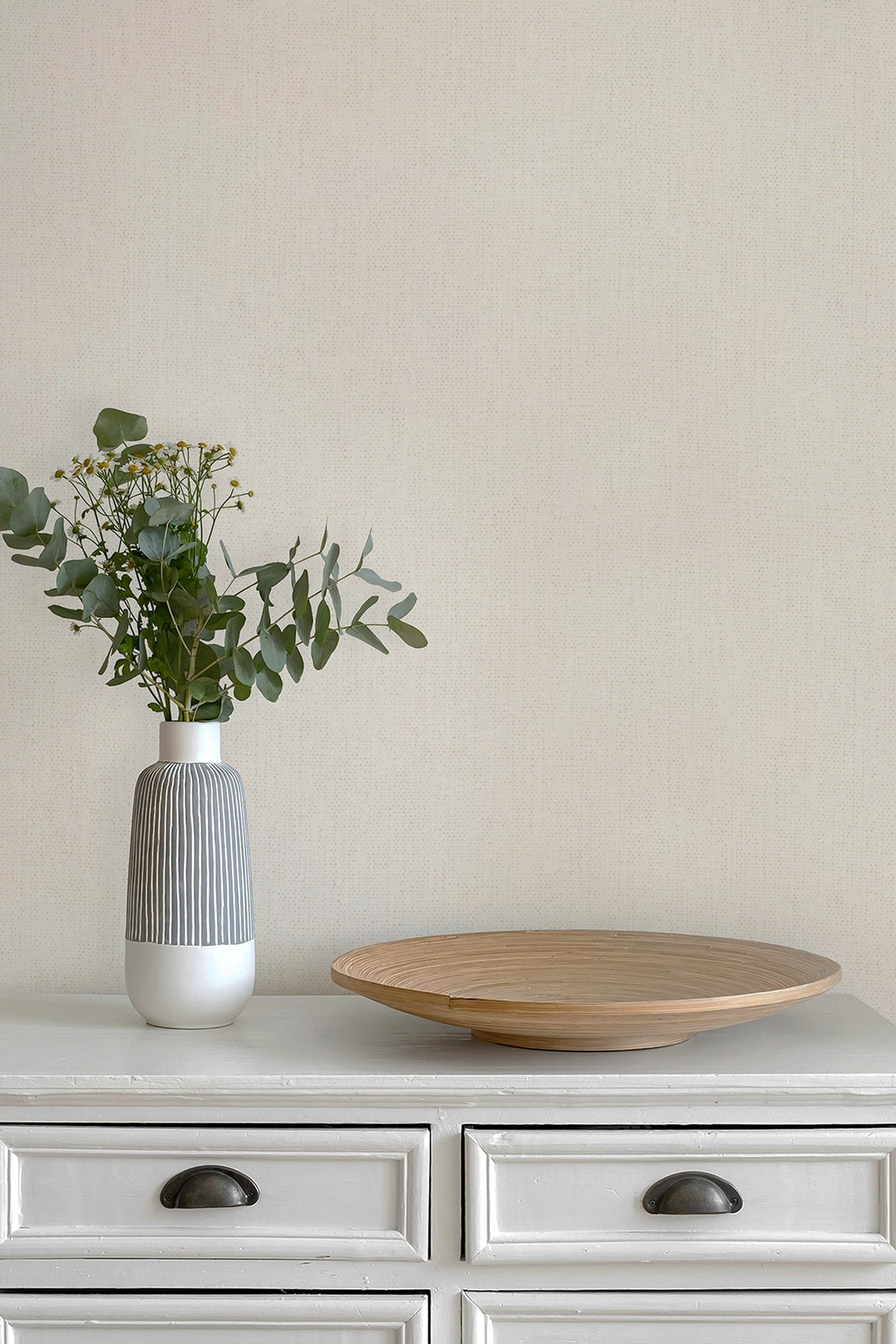 A modern ceramic vase with a gray ribbed body and white base holding green eucalyptus stems, placed on a white cabinet next to a round wooden tray against a wall in paperhaze wallpaper warm beige.