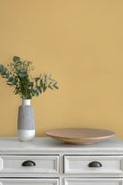 A styled interior scene with paperhze wallpaper in butter yellow, a white cabinet, a gray-and-white ribbed vase holding green eucalyptus stems, and a round light wood tray.