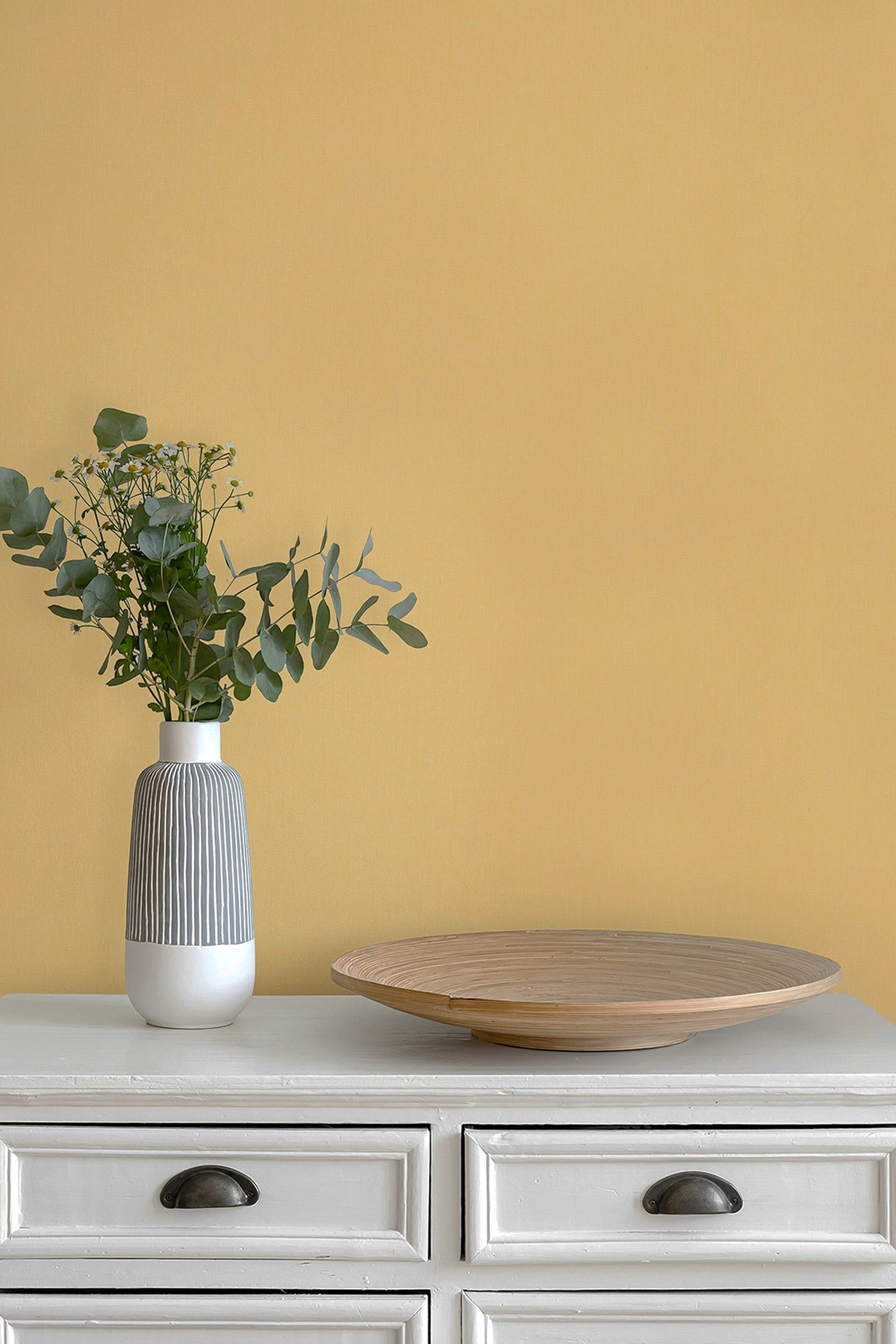 A styled interior scene with paperhze wallpaper in butter yellow, a white cabinet, a gray-and-white ribbed vase holding green eucalyptus stems, and a round light wood tray.