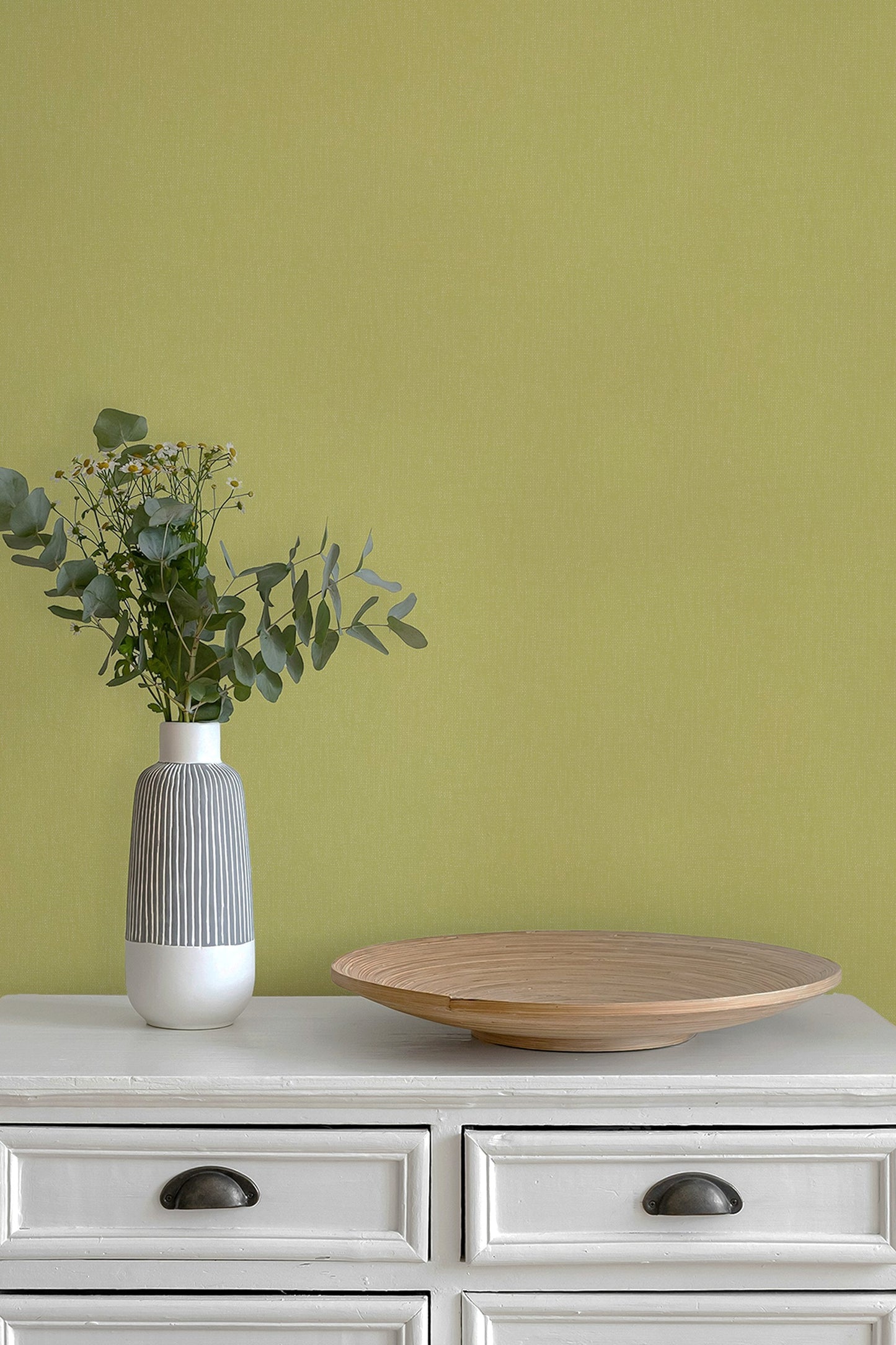 A styled interior scene with a white dresser against a paperhaze wallpapered wall in chartreuse. On top of the dresser sits a ribbed white and gray ceramic vase with green eucalyptus stems and a round light wood tray.