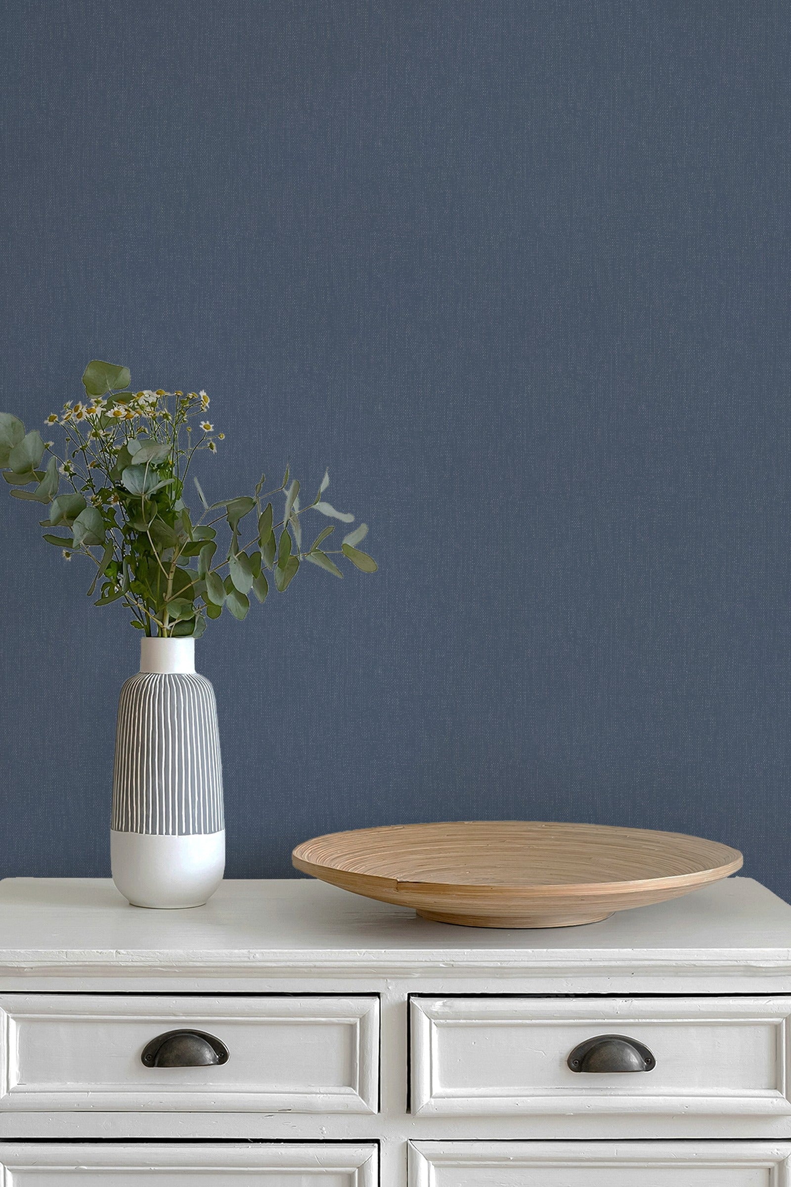 A styled interior scene with a white dresser against a paperhaze denim blue wallpaper, featuring a ribbed white and gray vase with greenery and a round wooden tray.