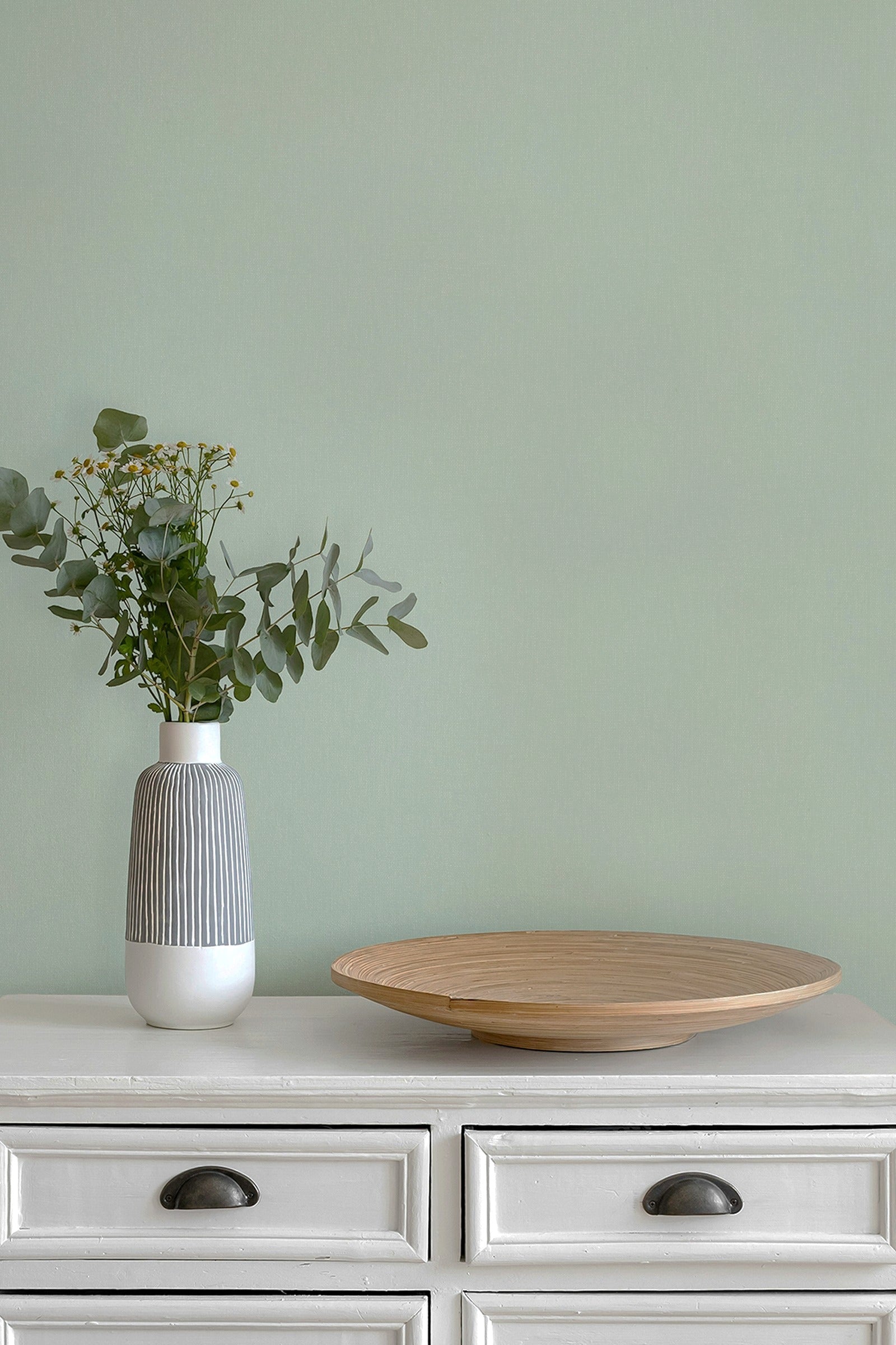 A styled interior scene with a paperhaze wallpaper in mint green, a white dresser, a gray-and-white ribbed vase with eucalyptus stems, and a round light wood tray.