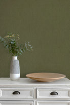 An olive green textured wallpaper shown on a wall behind a white wooden dresser with a gray and white ribbed vase holding greenery and a round wooden tray.