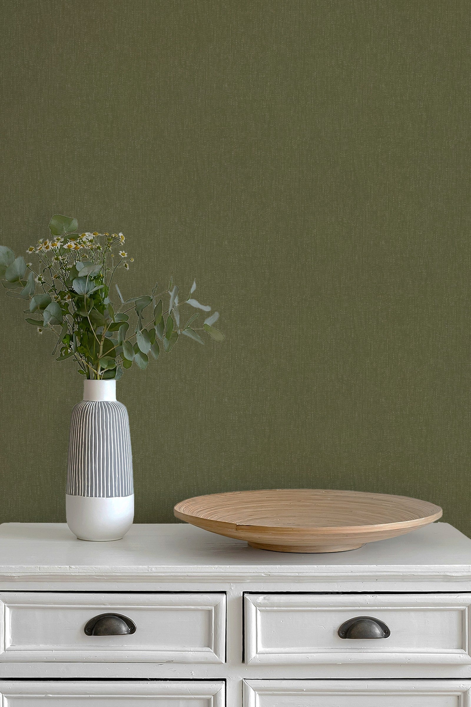 An olive green textured wallpaper shown on a wall behind a white wooden dresser with a gray and white ribbed vase holding greenery and a round wooden tray.