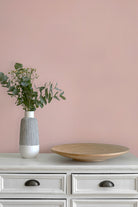 A styled interior scene with a shell pink paperhaze wallpapered wall, a white cabinet, a ribbed white and gray vase with green eucalyptus stems, and a round light wood tray.