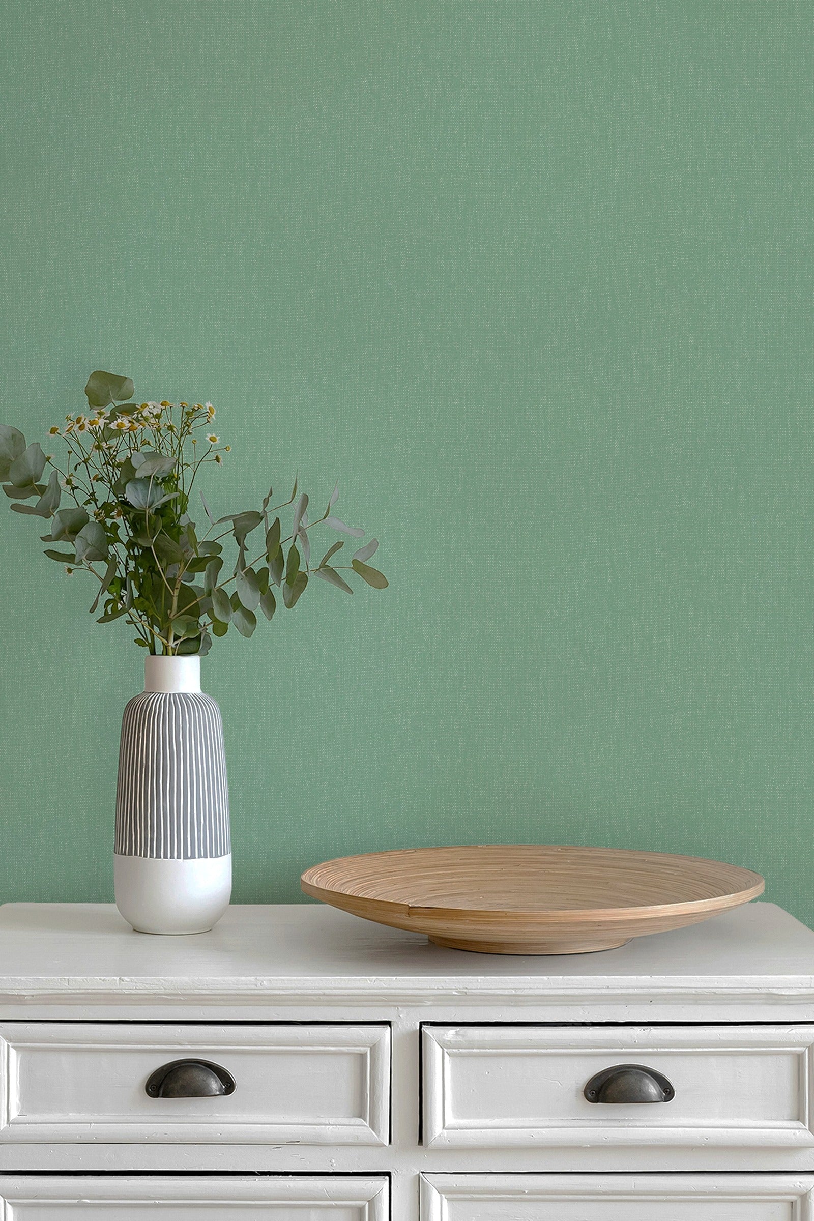A sage green textured wall behind a white wooden dresser with dark metal knobs, featuring a ribbed white and gray ceramic vase with eucalyptus stems on the left and a round light wood tray on the right.