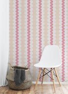 Pink and white wavy vertical stripes wallpaper shown in a styled room setting with a white molded plastic chair and a woven basket on a light wood floor.