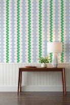 A wavy green stripe on oval scallop shapes with a blue grid background wallpaper its on top of a creamy white wainscot with a brown console table with a wooden bowl, vase of ferns, and a contemporary white ceramic table lamp.