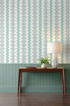 A styled interior scene showing a wall with vertical wavy stripes in mint green and white, above a teal wainscoted panel and dark wood console table. A white table lamp and a small potted plant sit on the table.